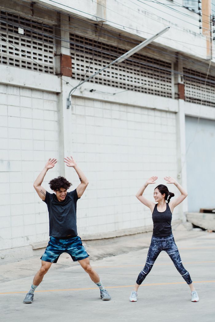 about-us-01 A man and woman performing jumping jacks outdoors by a building, focused on fitness.